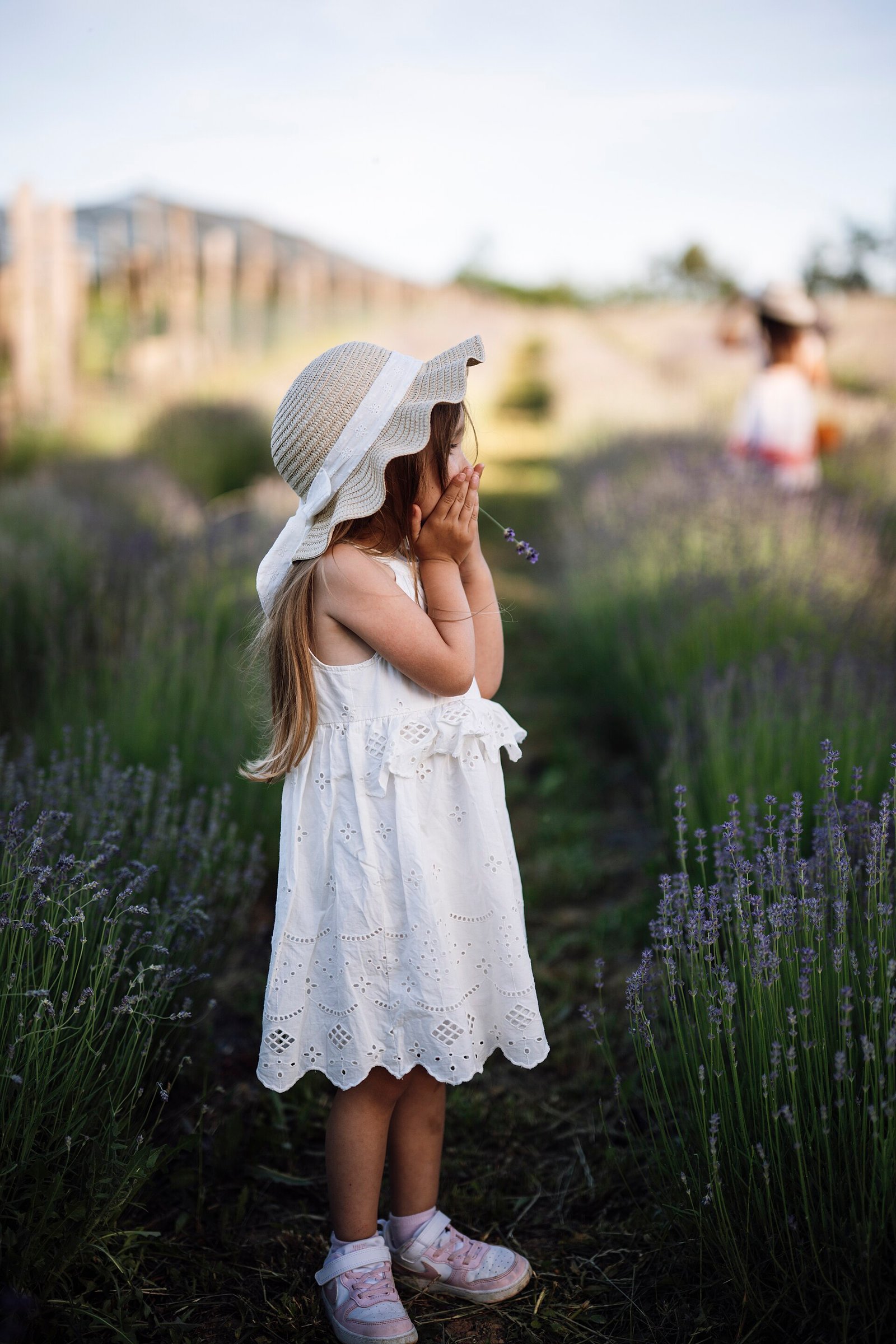 Menina de vestido claro explora campo de lavanda sob sol forte, protegida por chapéu de aba larga