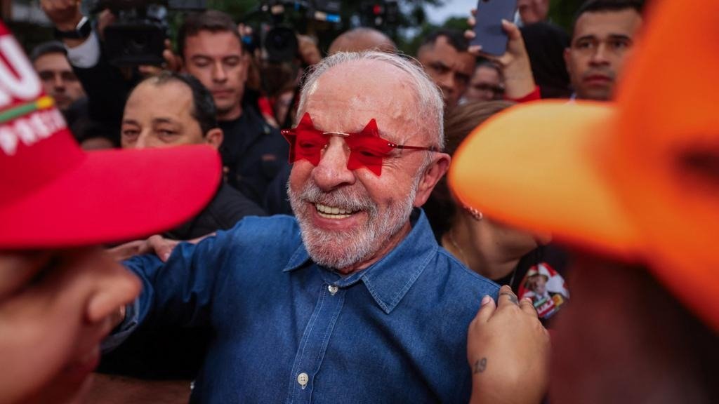 Lula sorrindo de óculos vermelhos em formato de estrela, cercado por apoiadores e jornalistas