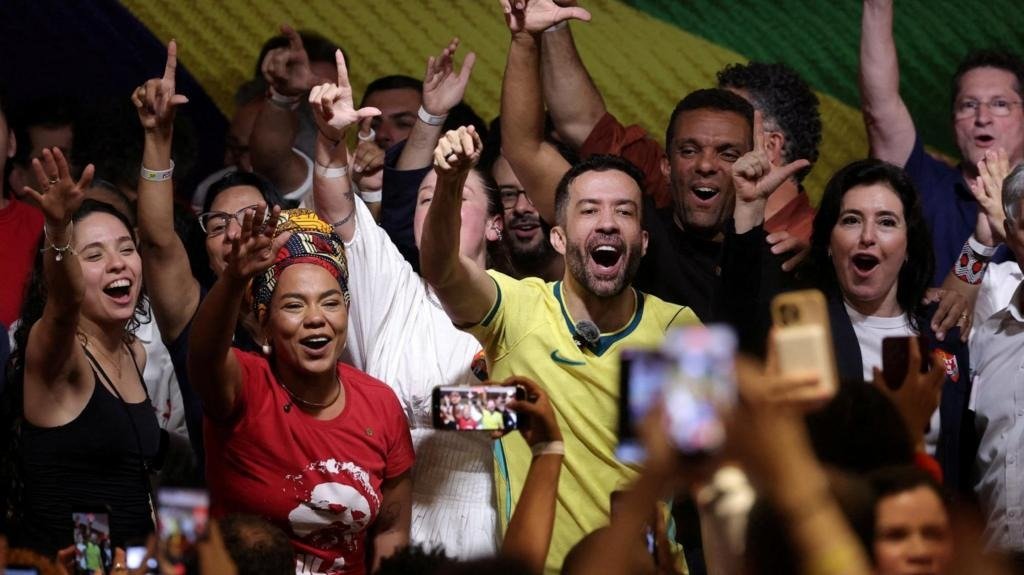 Multidão em celebração com bandeira do Brasil ao fundo e apoiador de camisa amarela ao centro